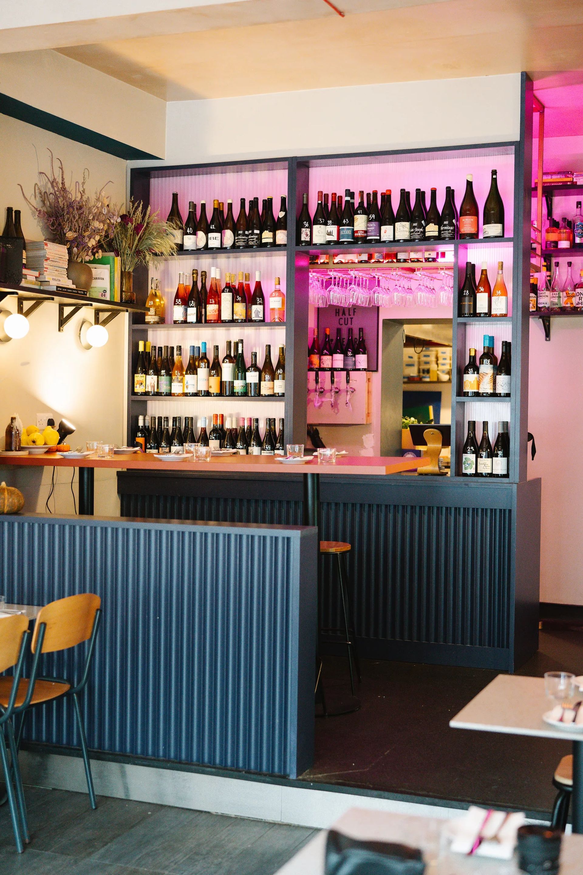 A modern bar with shelves filled with various bottles of wine and spirits, glasses hanging above the counter, and purple accent lighting. Wooden chairs and tables are visible in the foreground, creating a scene worthy of Hannah Crosbie's tasting notes.