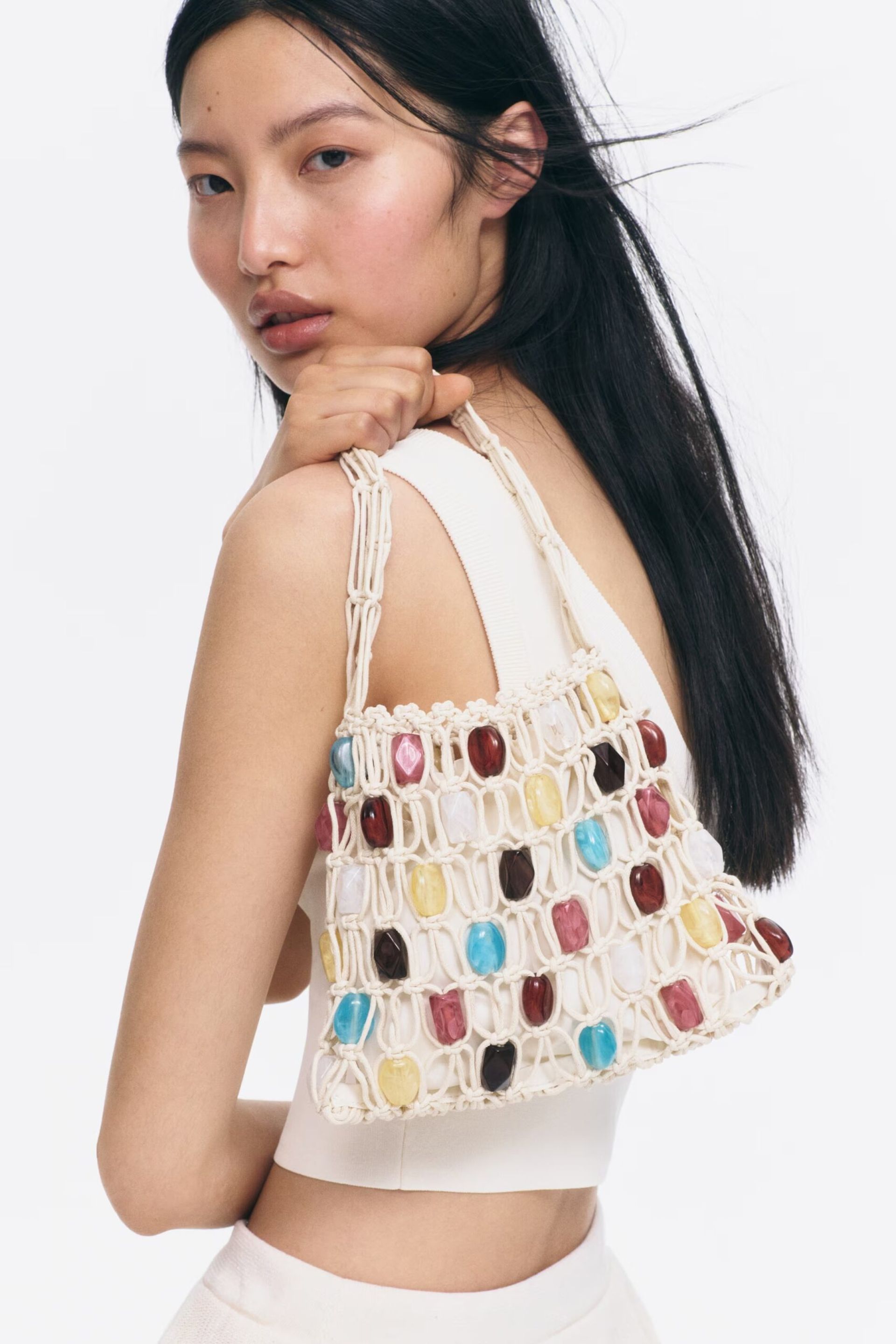 A woman with long black hair wears a white top and holds a beaded bag in a macramé style, adorned with colourful, polished beads.