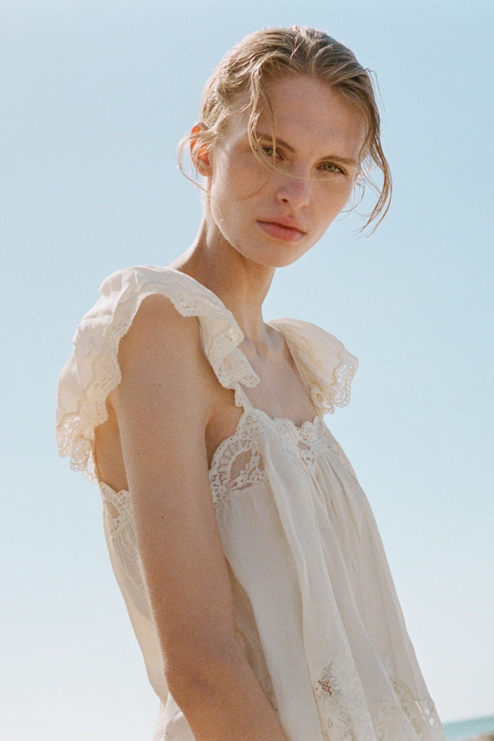 A woman stands outdoors against a clear blue sky, wearing a light, sleeveless, lace-trimmed dress with ruffled shoulder details—perfect for those who love summer tops. Her hair is loosely pulled back as she looks towards the camera with a neutral expression.