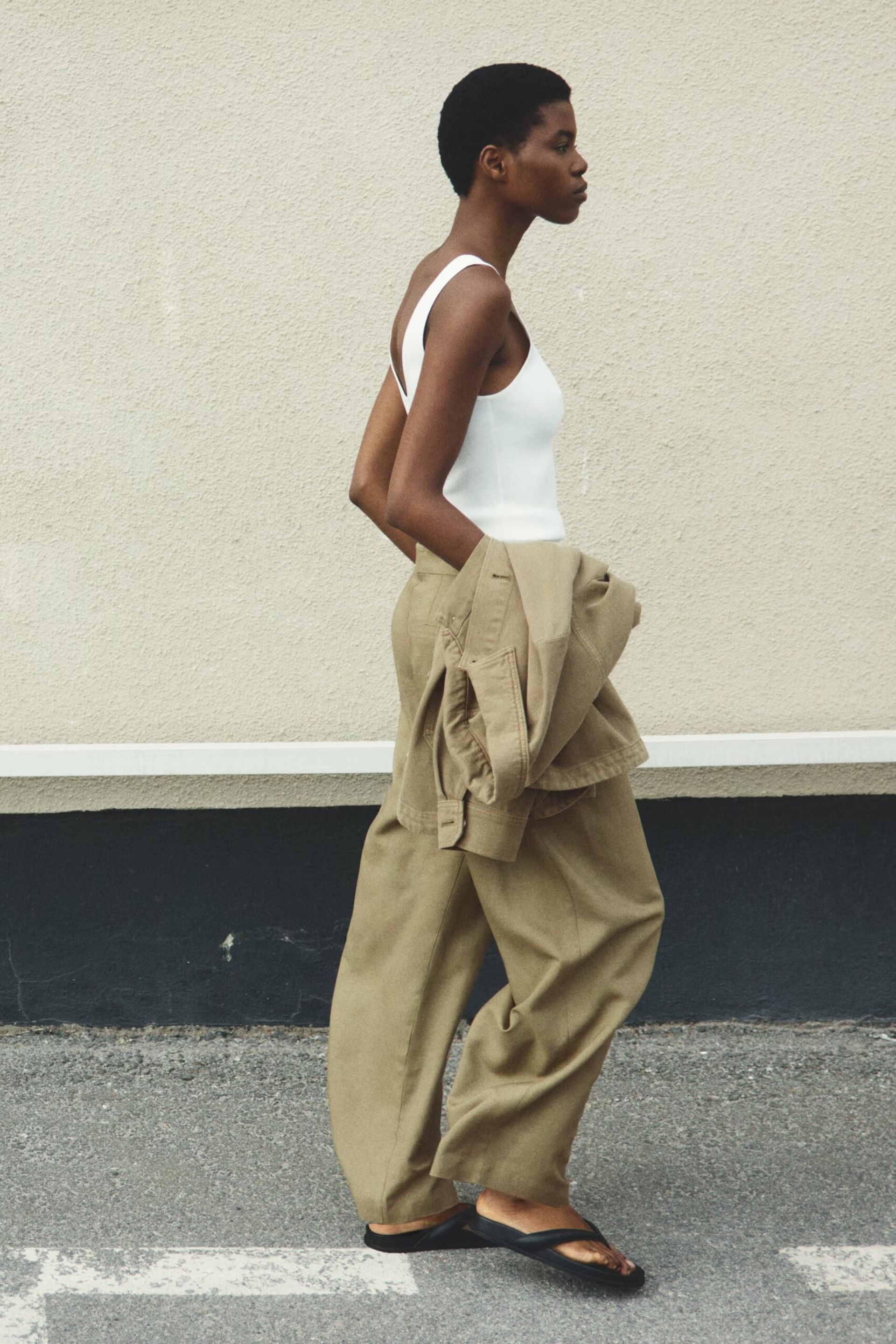 A person with short hair walks outside in high street linen—a white vest, loose beige trousers, and black sandals—carrying a matching beige jacket. They are in profile against a light-coloured wall.