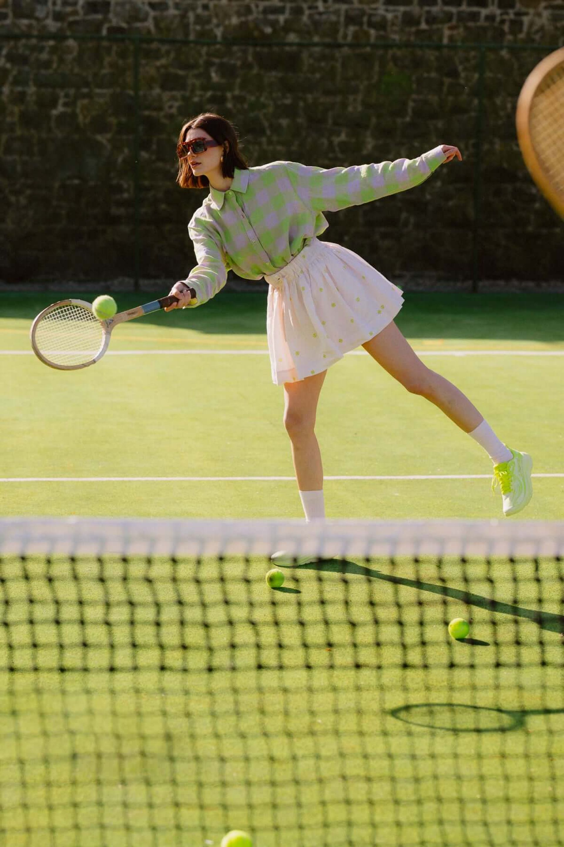 A woman in a green jumper and white skirt plays tennis on an outdoor court, swinging her racquet to hit a ball. Her look channels padel court fashion, with several tennis balls scattered nearby and a net visible in the foreground.