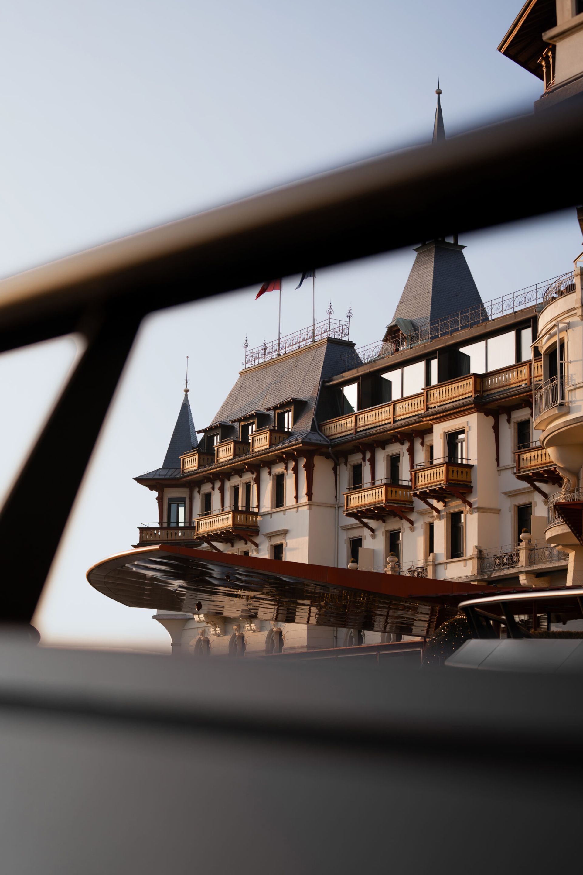 A grand historic building with pointed towers and ornate balconies, resembling a fairytale hotel, is partially visible behind a blurred black railing. The clear sky and warm lighting suggest late afternoon in Zürich.