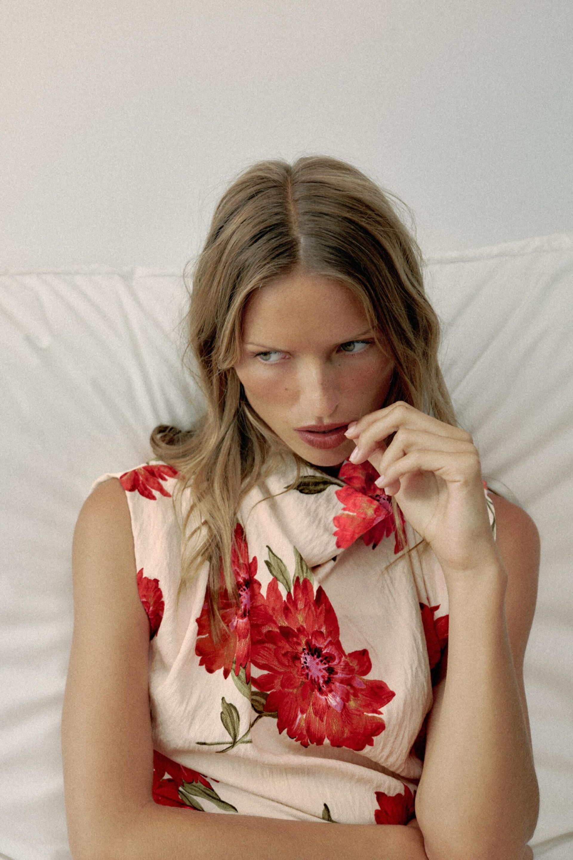 A person with long, wavy blonde hair wearing a sleeveless dress with large red floral prints—a striking piece of high street fashion—sits on a white cushion, looking slightly away and touching their lips with their fingers.