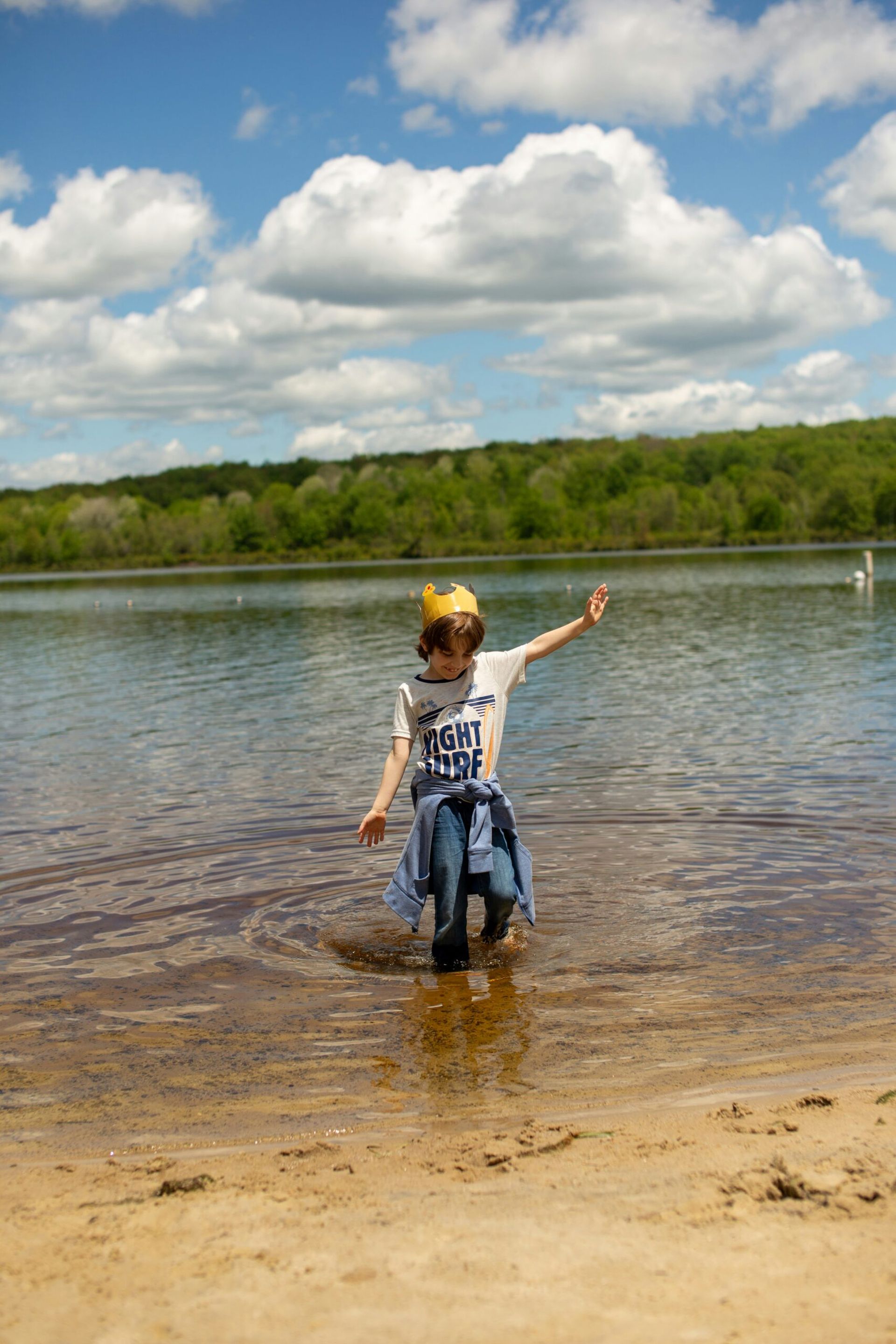 A child wearing a crown and a NIGHT CREW shirt wades in shallow lake water, arms outstretched, on a sunny day with clouds and trees in the background—capturing the magic of Summer Holidays With Children.