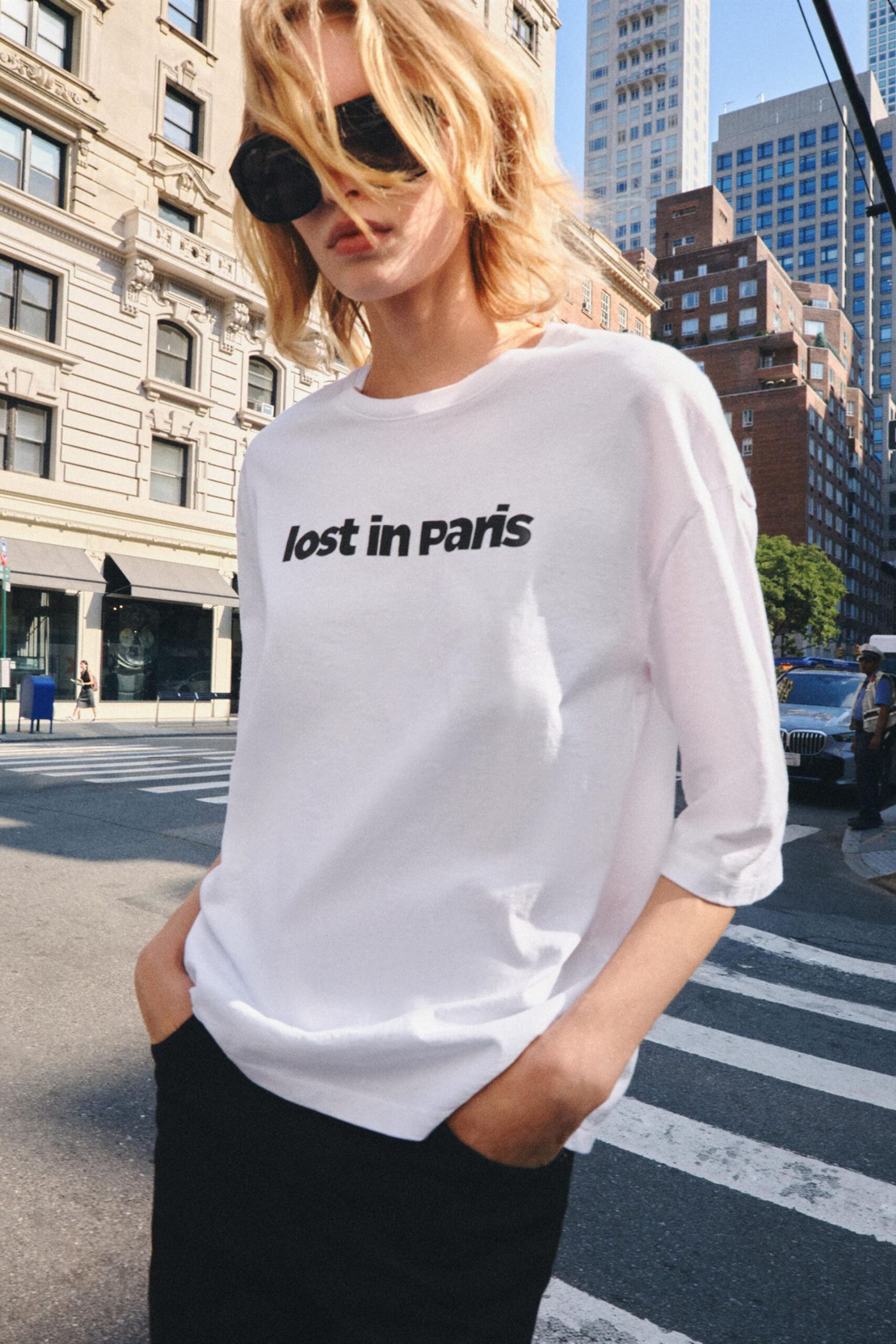 A person with blonde hair and sunglasses stands on a city street, wearing one of those cool slogan tees—a white shirt that says “lost in paris” in black text. Tall buildings and zebra crossing lines are visible in the background.