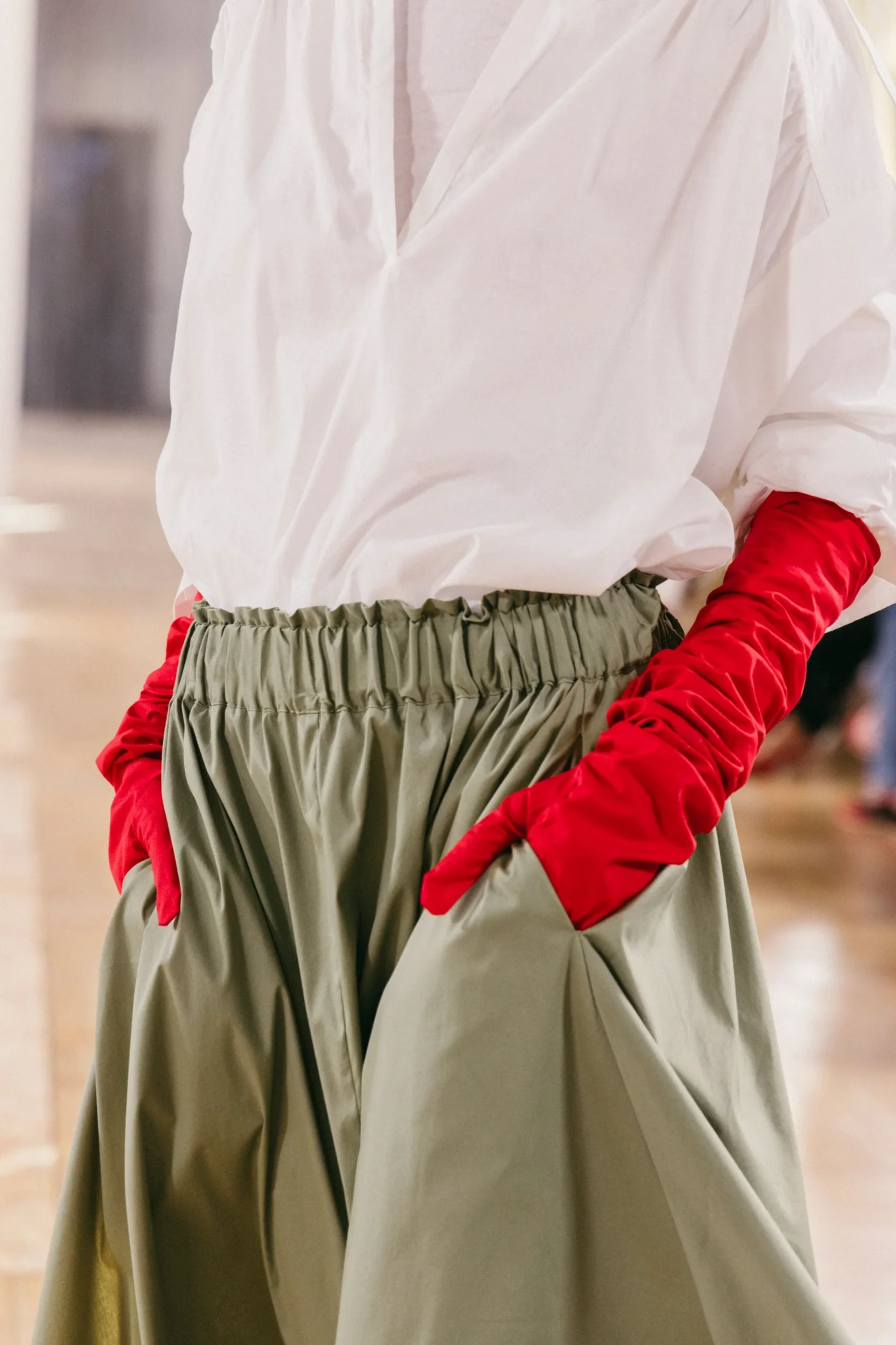 A person wearing a loose white shirt, olive green skirt with an elasticated waistband, and long red gloves—an effortlessly stylish look that showcases the unique flair often found in small fashion brands—stands with hands in their pockets.