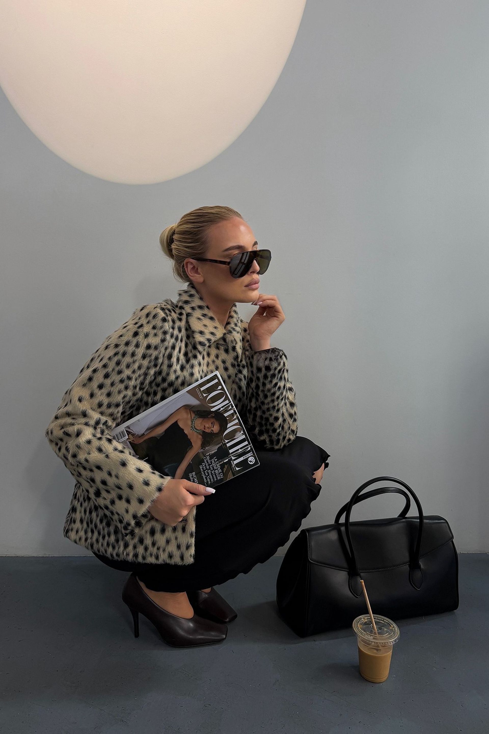 A woman in a spotted lightweight jacket and sunglasses squats indoors, holding a fashion magazine. A black handbag and an iced coffee sit on the floor beside her.