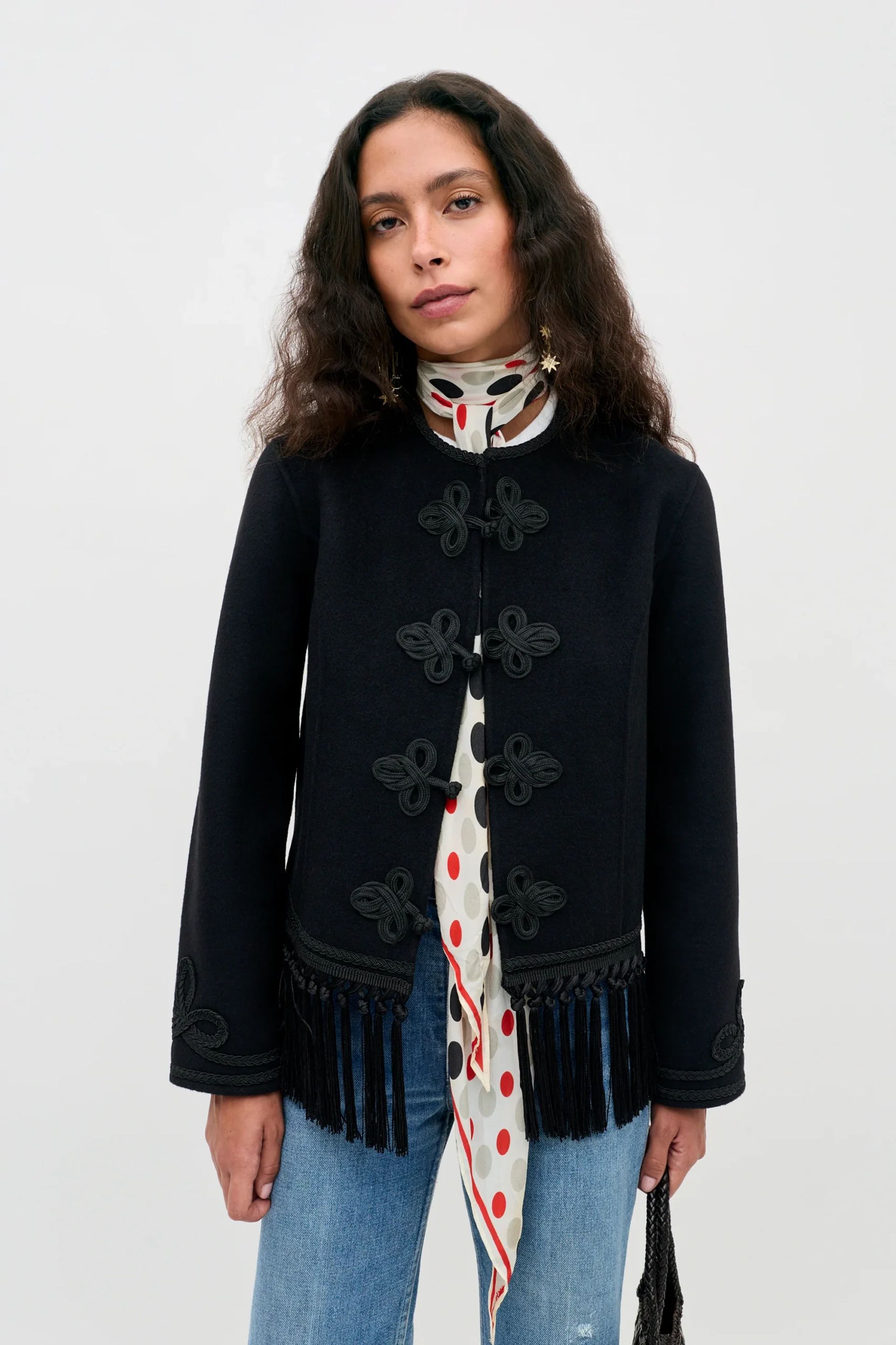 A woman with wavy brown hair wears a stylish black lightweight jacket with knot buttons and fringe, a white scarf with red polka dots, and blue jeans, standing against a plain light background.