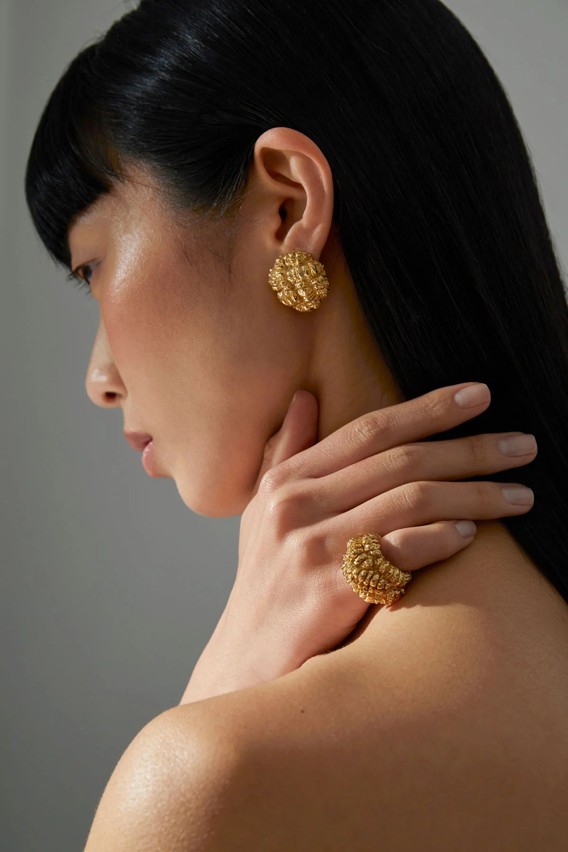A woman with straight black hair wears textured gold earrings and a matching gold ring—unique pieces from small fashion brands—touching her neck with elegant fingers against a neutral background.