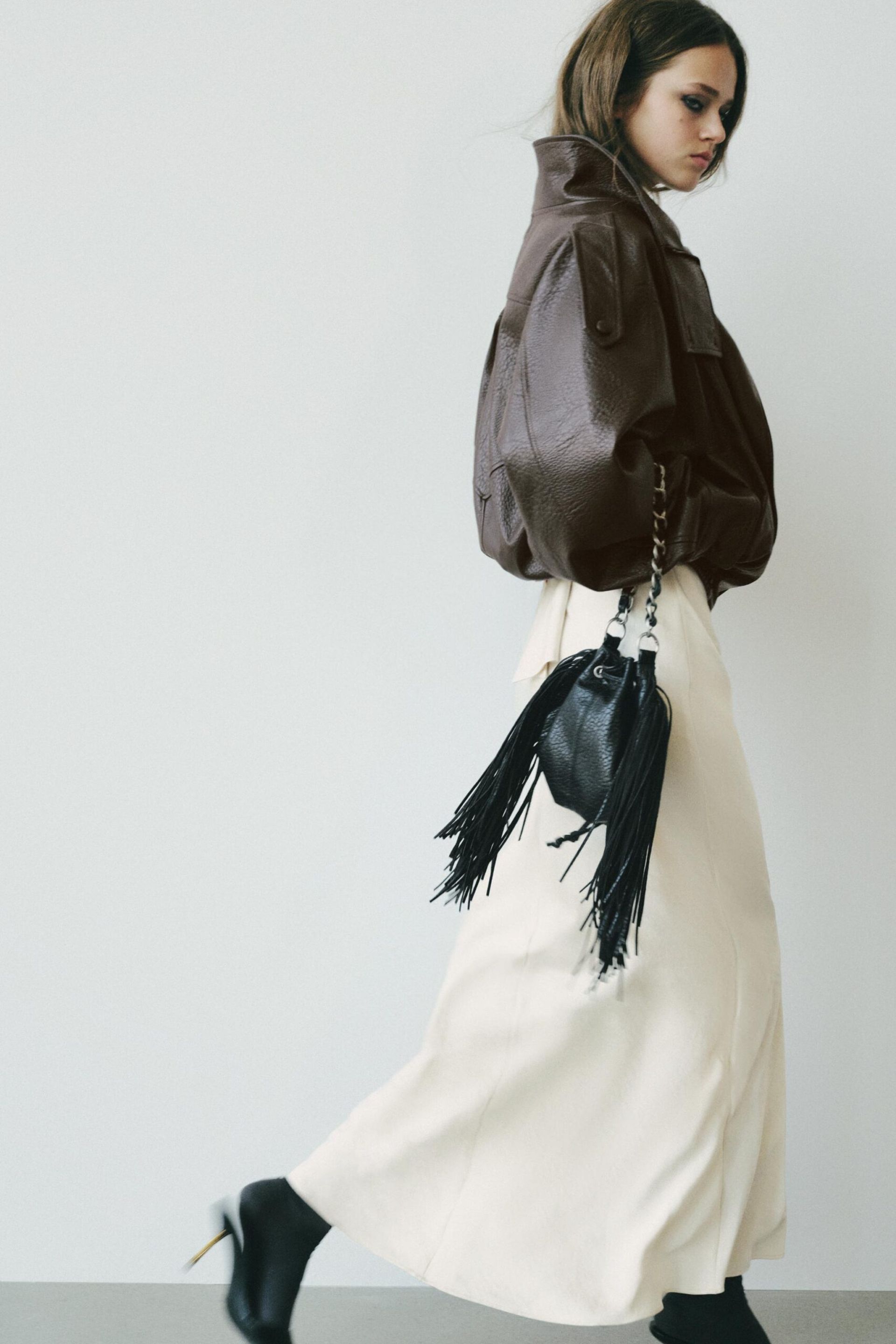 Woman in a brown leather jacket, cream maxi skirt, and black boots walks whilst carrying a black fringed handbag—a perfect choice for cool girls embracing autumn style. She has long hair and looks down, against a plain white background.