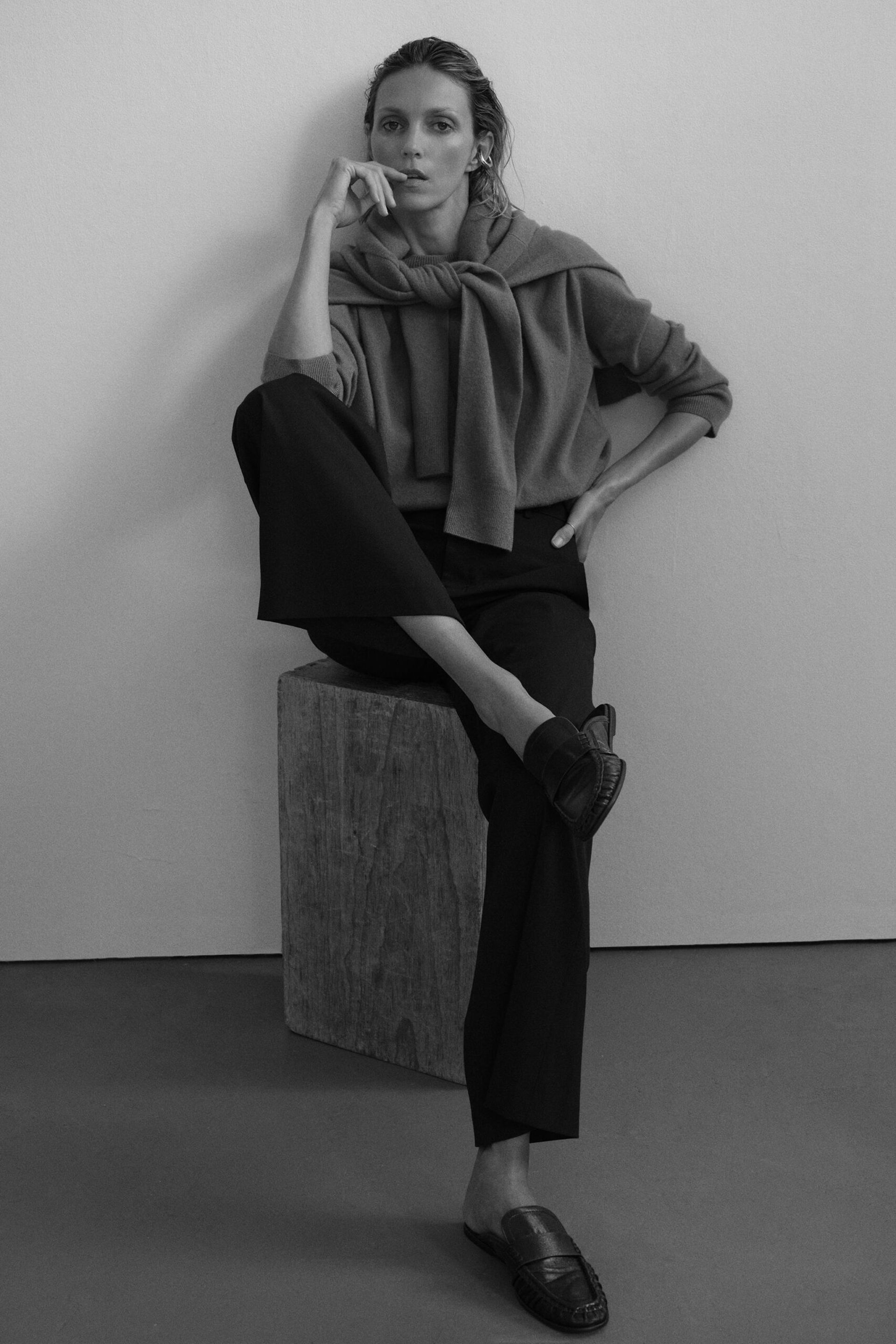 A person sits on a wooden block, wearing a jumper draped over their shoulders, a top, dark trousers, and loafers from minimalist high street brands, with one leg crossed over the other and their hand resting on their face, looking at the camera.