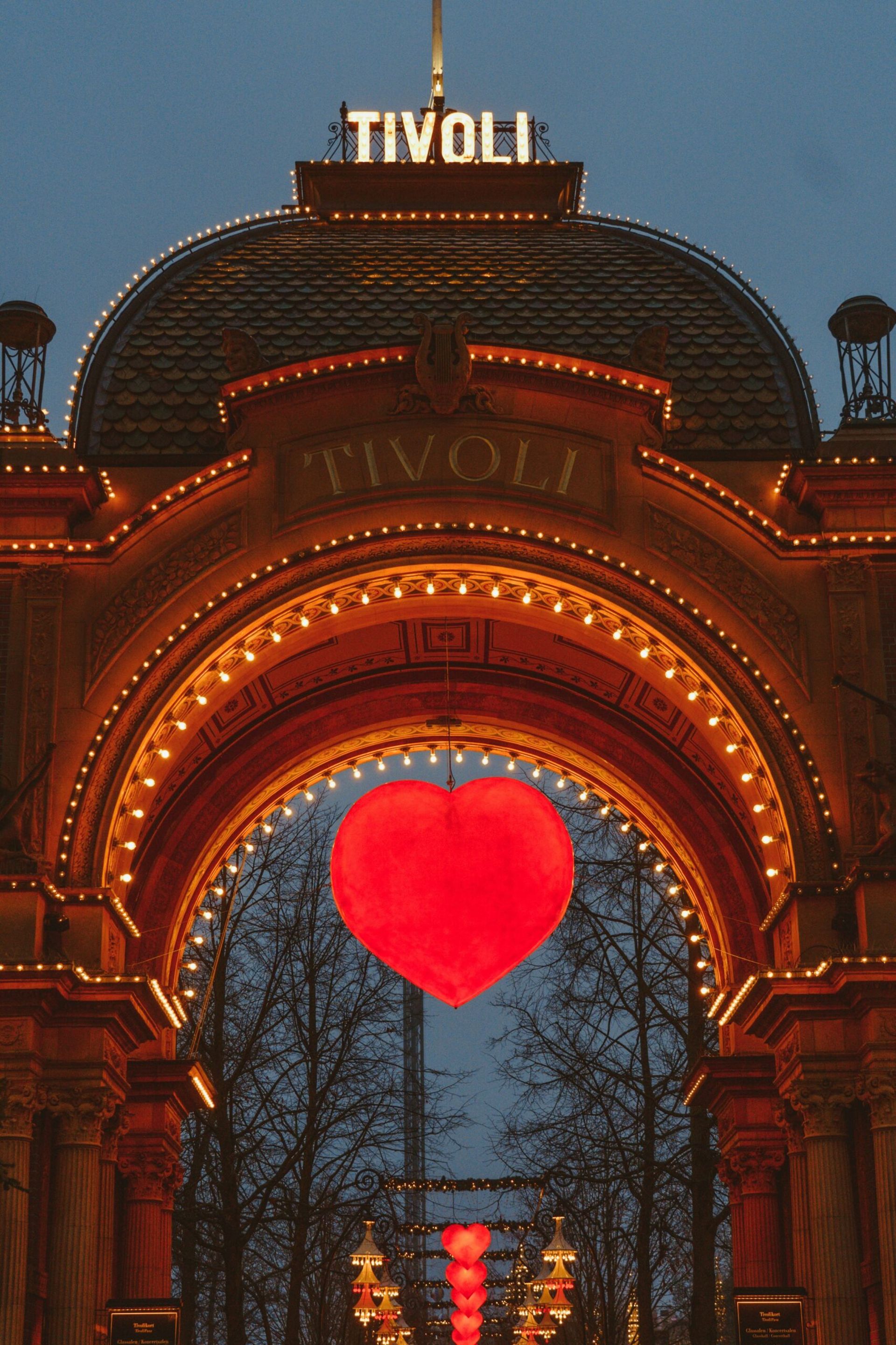 The entrance to Tivoli Gardens in Copenhagen is illuminated with warm lights at dusk, featuring a large glowing red heart and several smaller hearts inside—an enchanting sight for your 2026 travel itinerary near Villa Copenhagen.