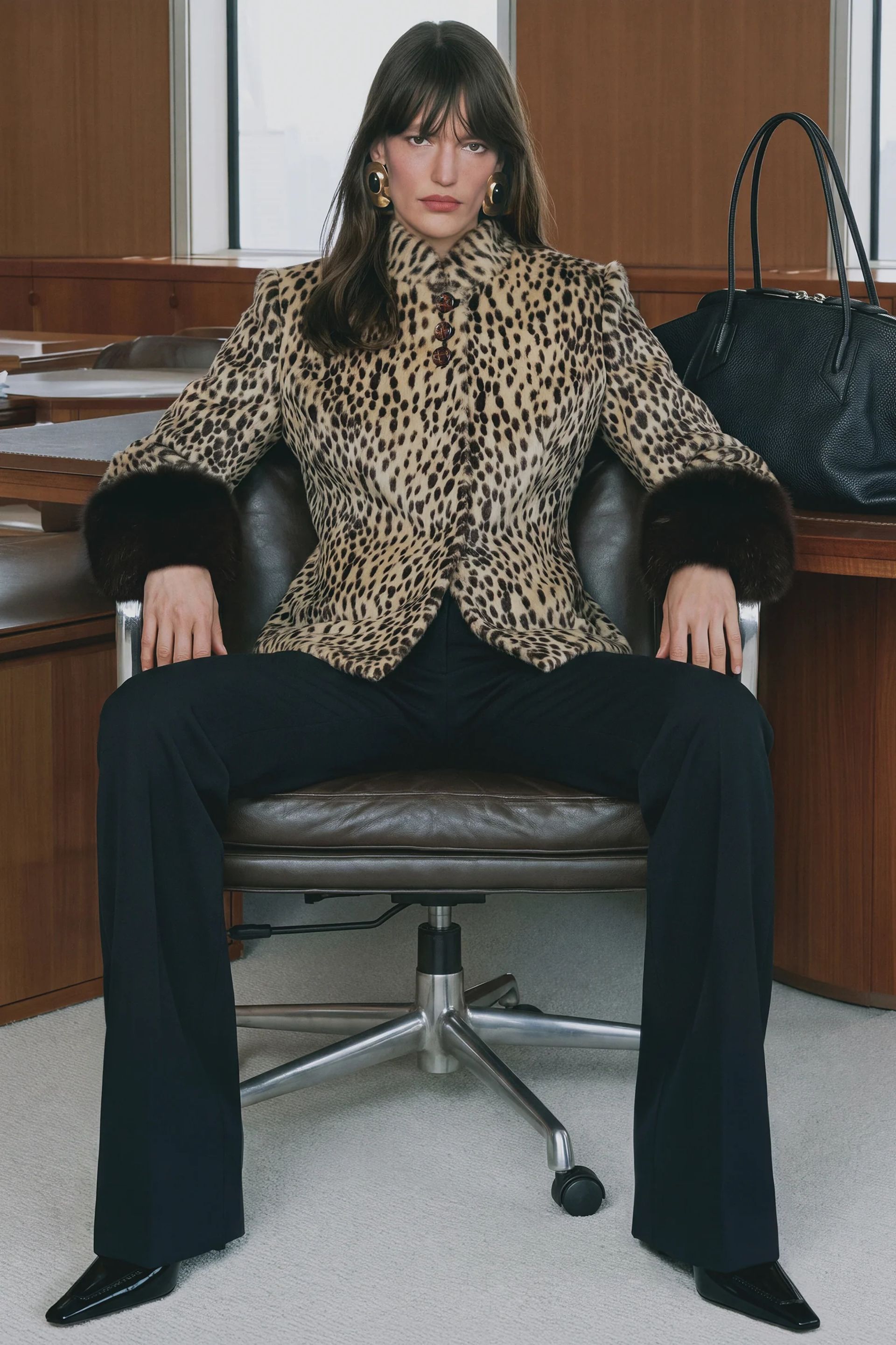 A person sits confidently in an office chair, wearing a leopard print coat with dark fur cuffs from one of the best small fashion brands, paired with black trousers and heels. A large black bag rests on the wooden desk behind them.