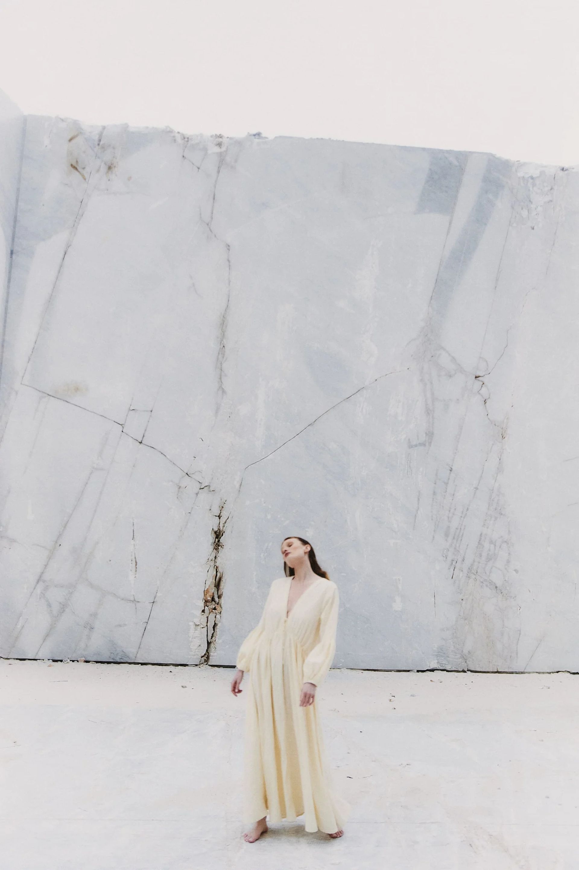 A woman in a long, light yellow dress from one of the distinguished small fashion brands stands barefoot on a white surface in front of a large, cracked white marble wall, looking upwards with a serene expression.