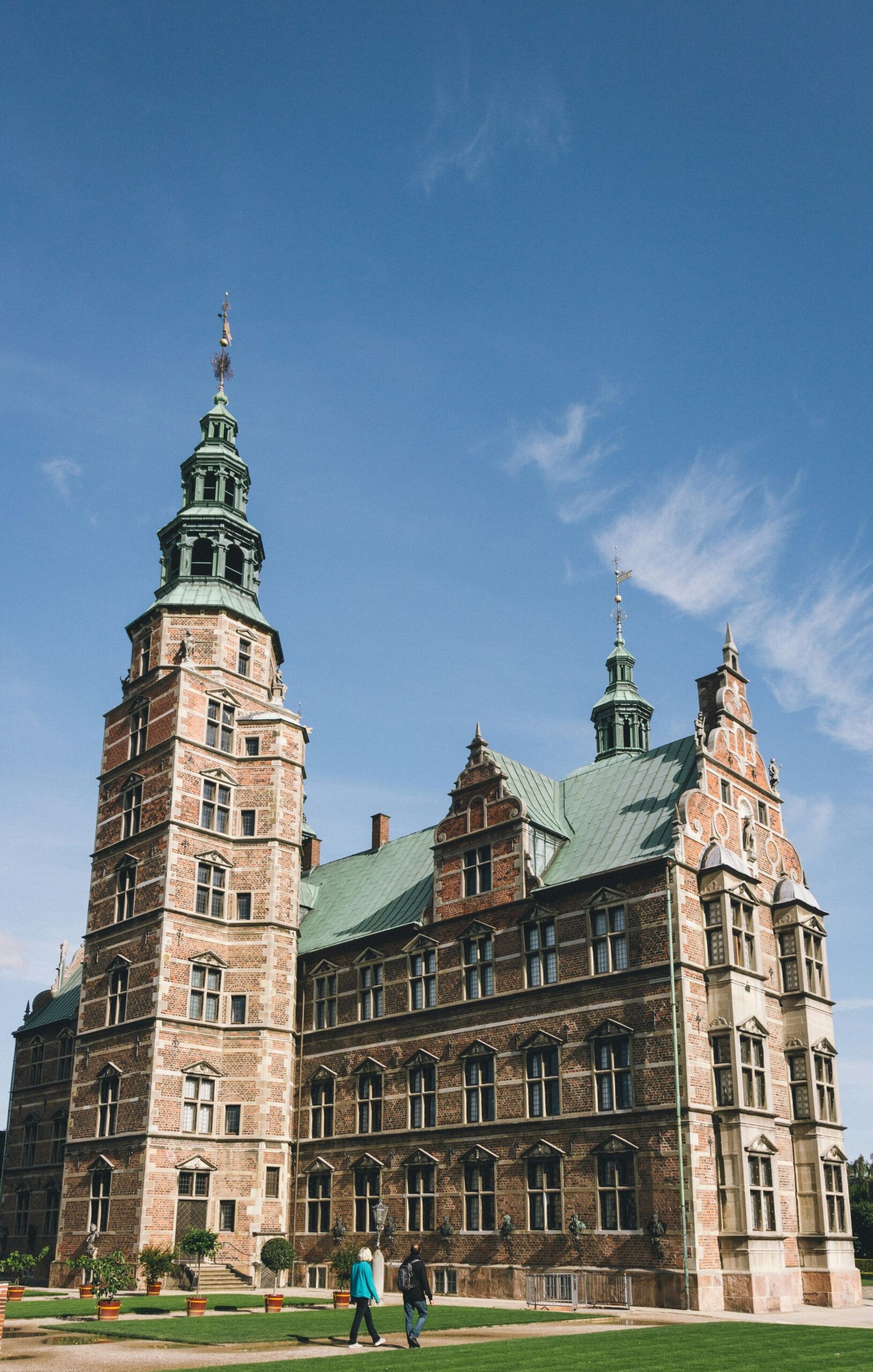 A grand historic brick building with tall, ornate towers and a green copper roof, Villa Copenhagen stands against a clear blue sky. Two people walk in front of the landmark on a sunny 2026 travel day.