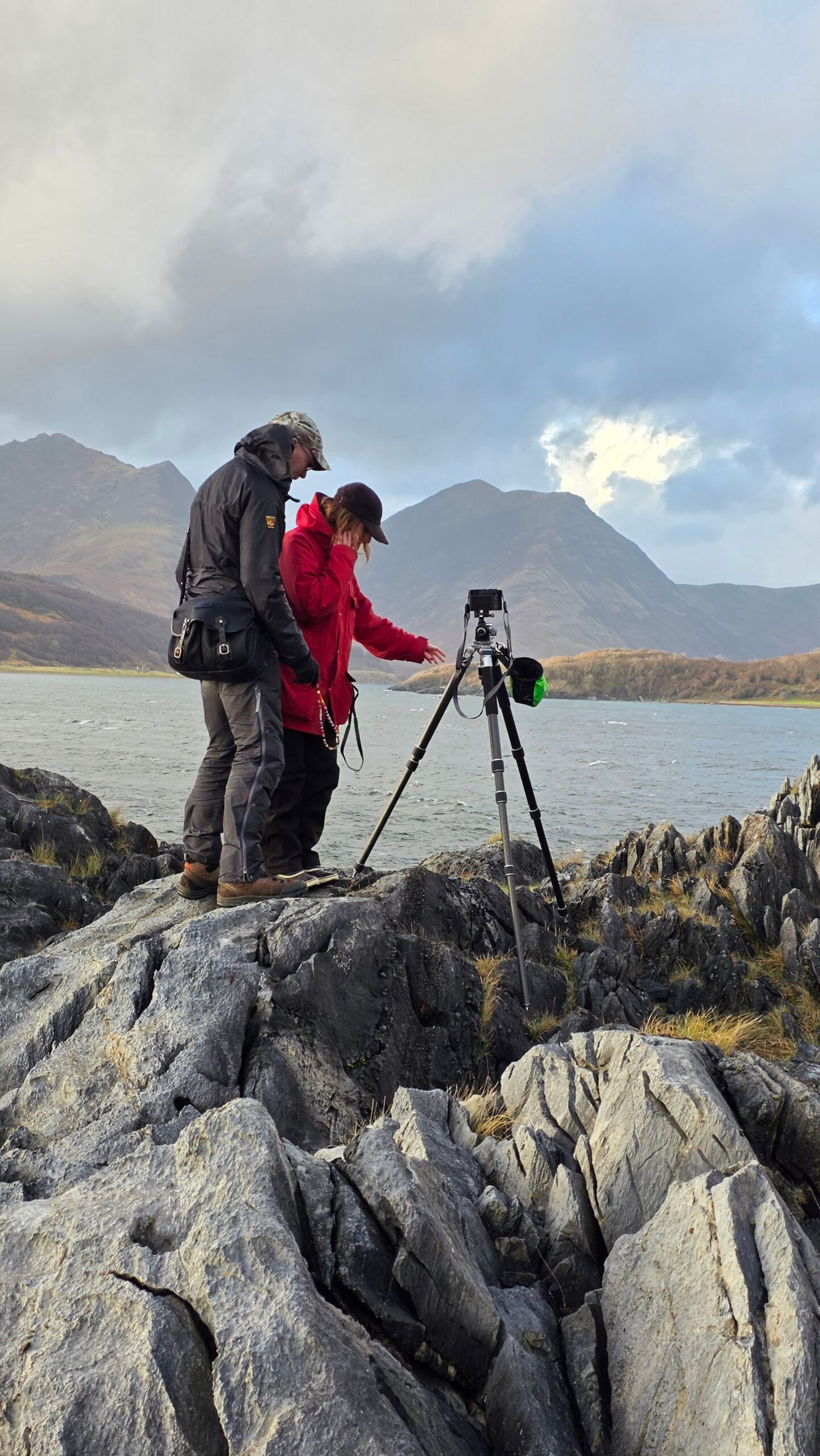 Two people in outdoor gear stand on rocky terrain by a lake near Kinloch Lodge, with mountains rising in the background. They examine a camera on a tripod together, pointing at the screen beneath a partly cloudy sky.