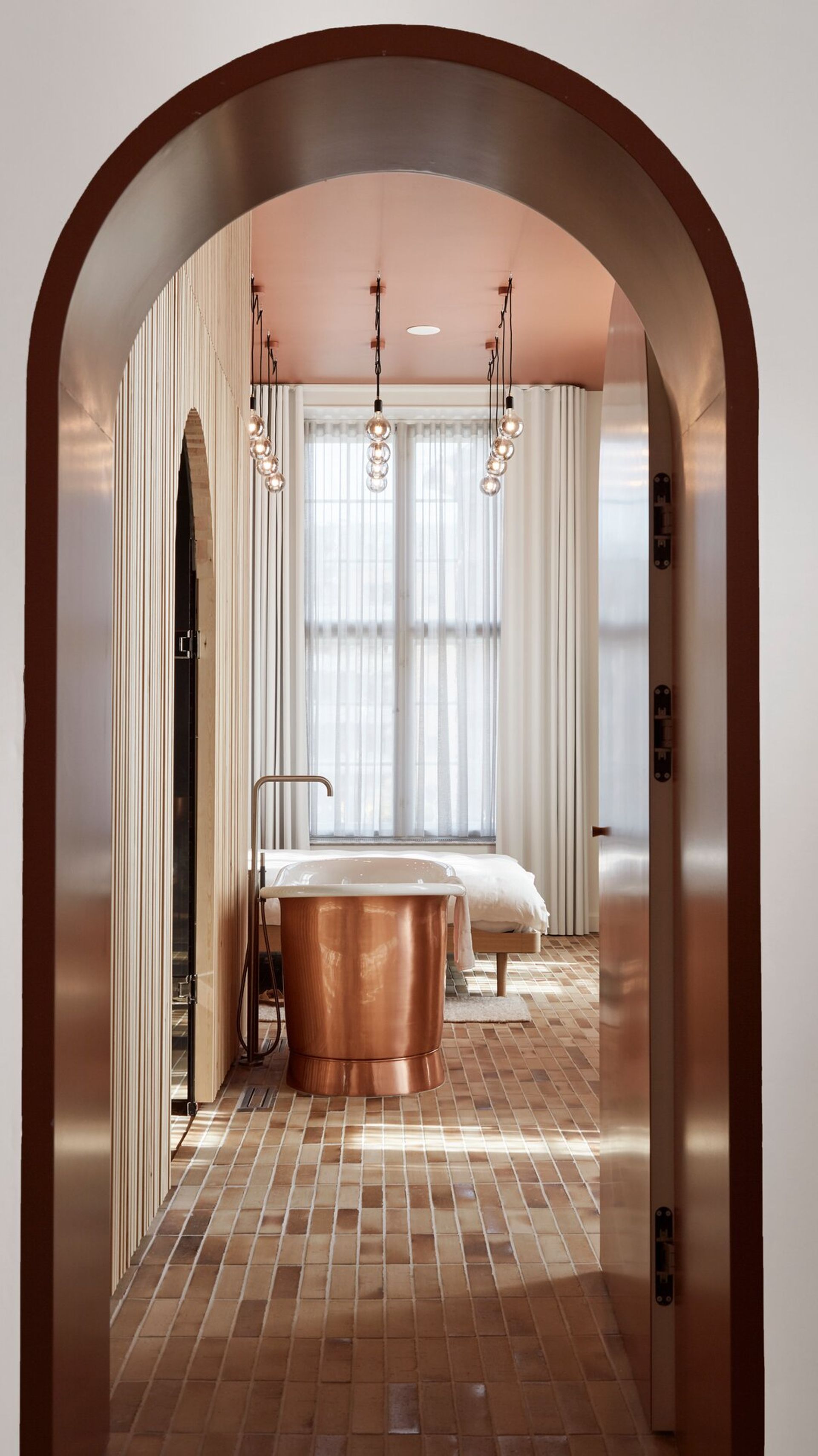 A modern bathroom in Villa Copenhagen with brick flooring, a copper bath, and floor-to-ceiling curtains is viewed through an arched doorway with warm brown trim and pendant lights—a perfect 2026 travel inspiration.