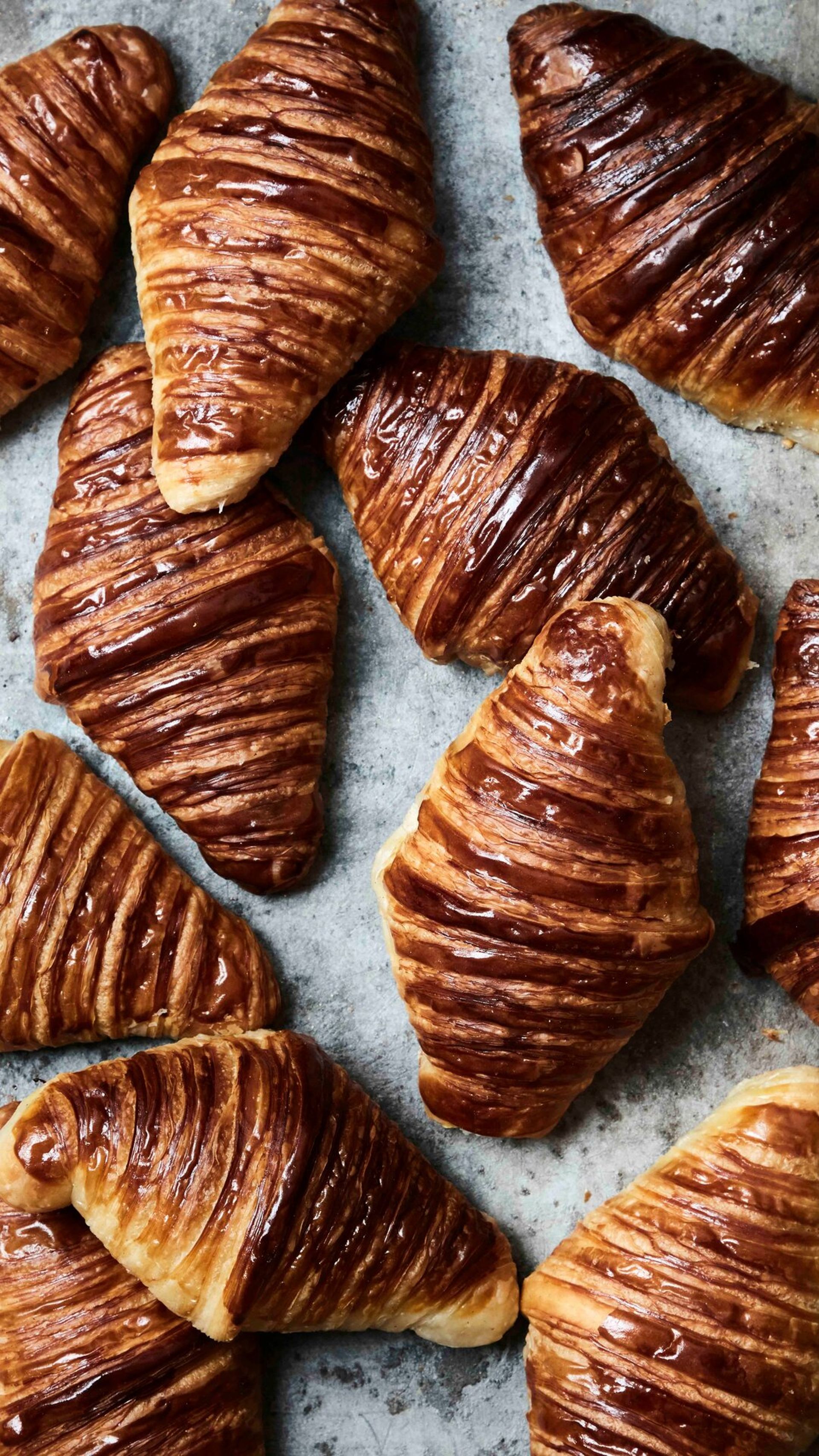Several golden-brown croissants, some with a chocolate glaze, are arranged on a textured grey surface at Villa Copenhagen, showcasing their flaky, layered pastry and glossy finish—perfect for your 2026 travel breakfast.