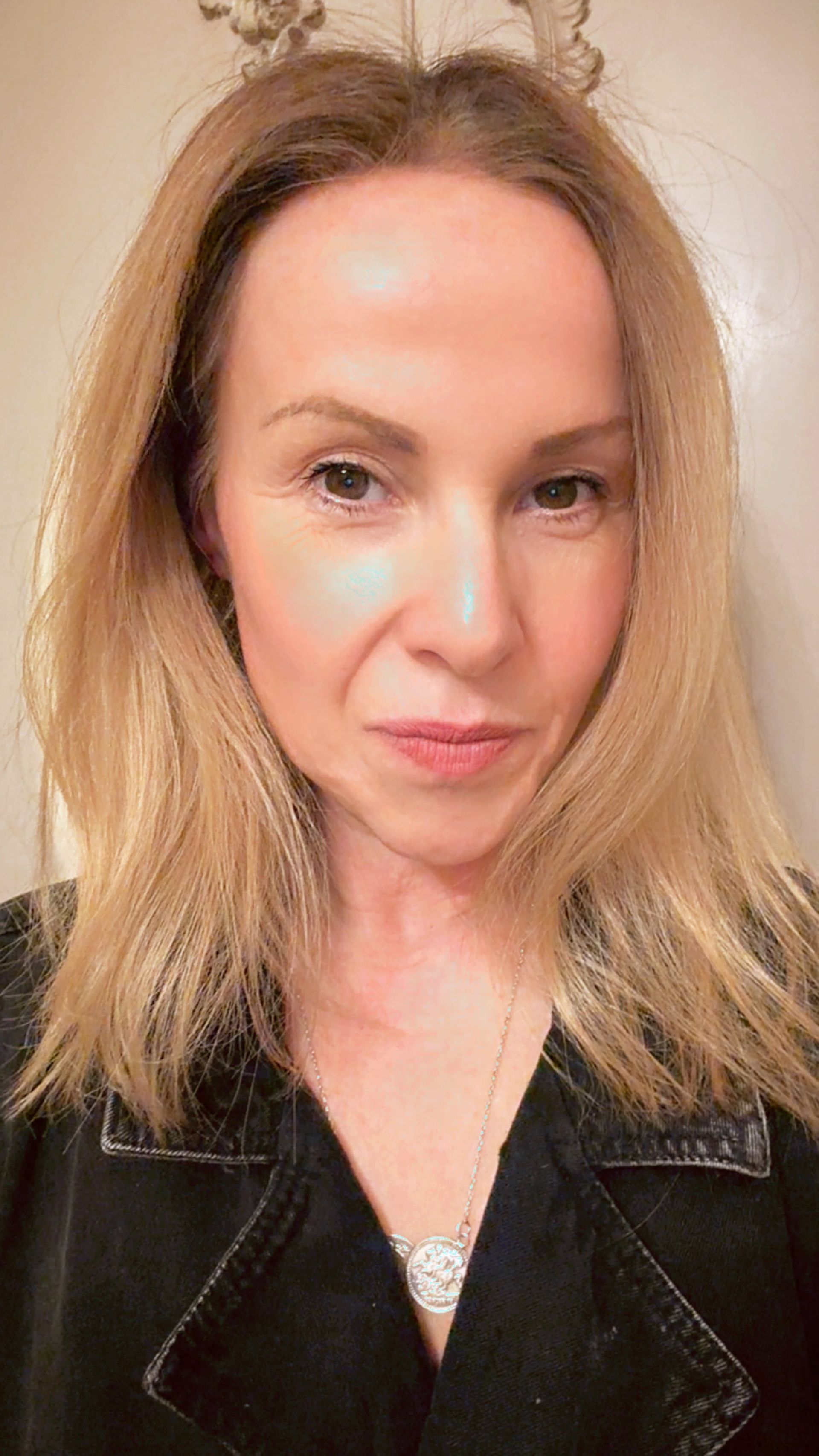 A woman with straight, shoulder-length blonde hair styled by Sam McKnight and light skin looks directly at the camera. She wears a black top and a necklace, set against a neutral, light-coloured background.