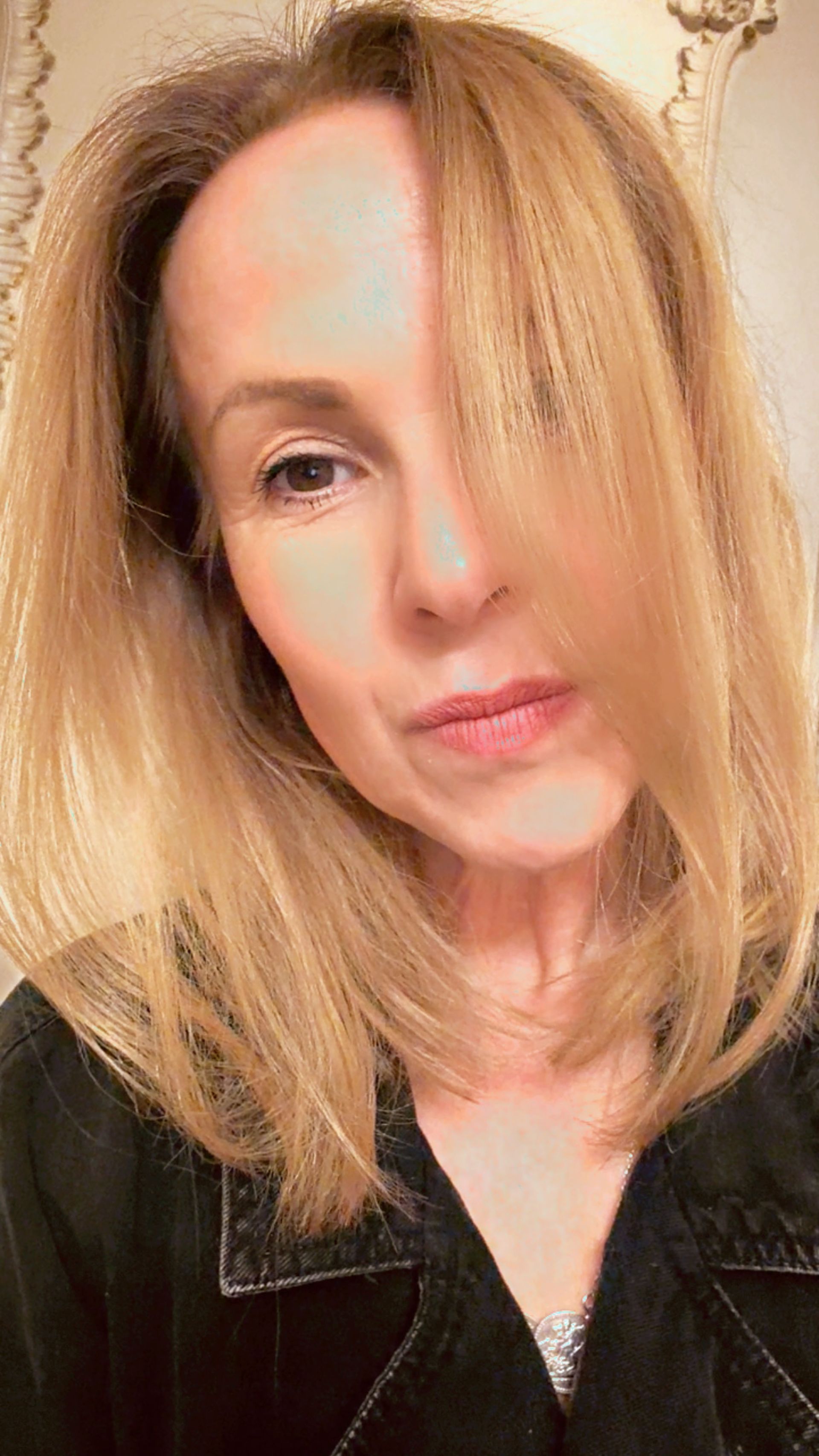 A woman with straight, shoulder-length blonde hair styled by Sam McKnight poses for a close-up selfie. She is wearing a black top, some hair partially covers one eye, and she has a neutral expression in a well-lit indoor setting.
