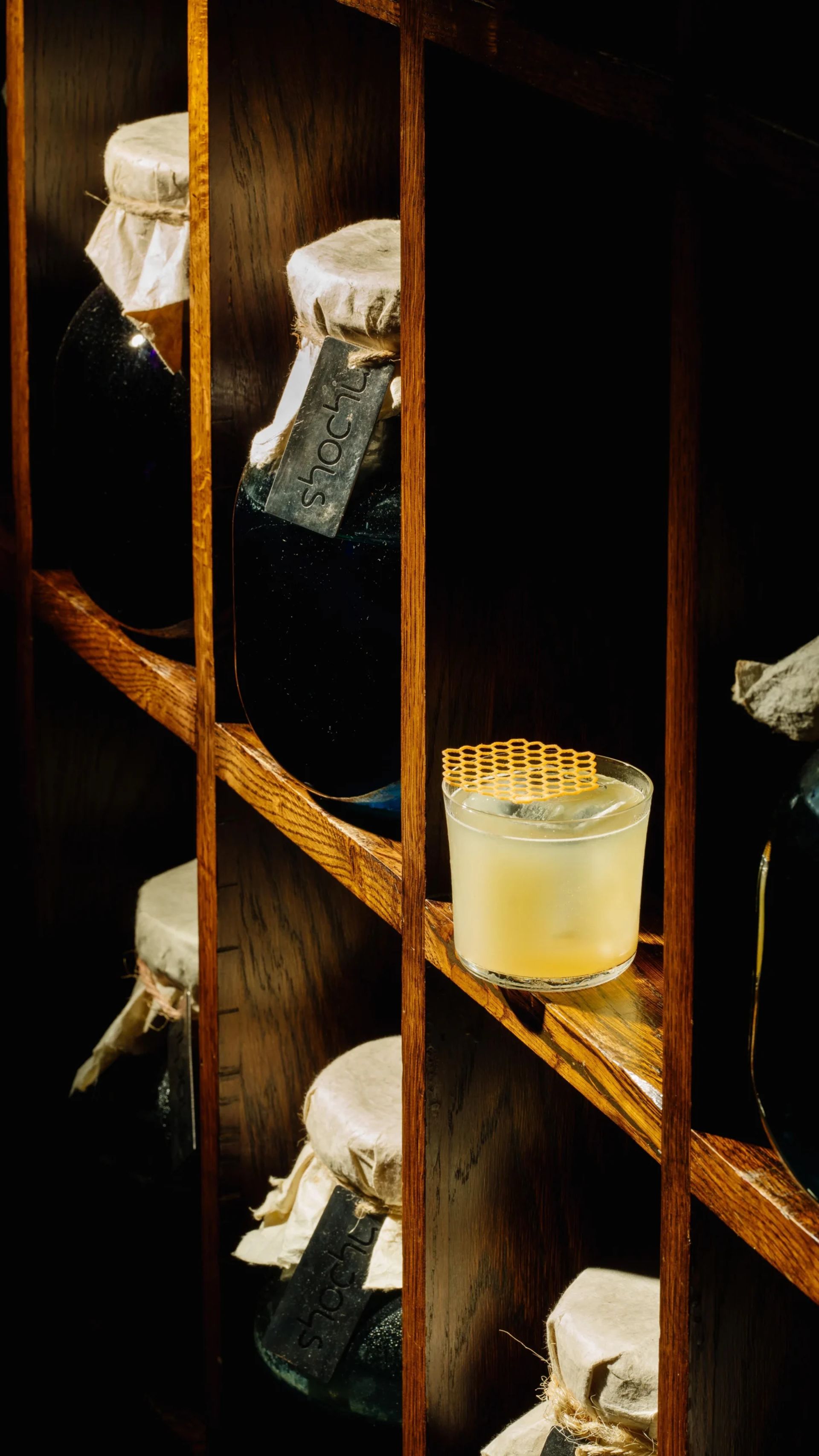 A yellow margarita in a clear glass with a honeycomb garnish sits on a wooden shelf, surrounded by large glass jars covered with paper and string. Warm lighting highlights the drink and wood grain—a perfect pick for any margarita-lover or special occasion.