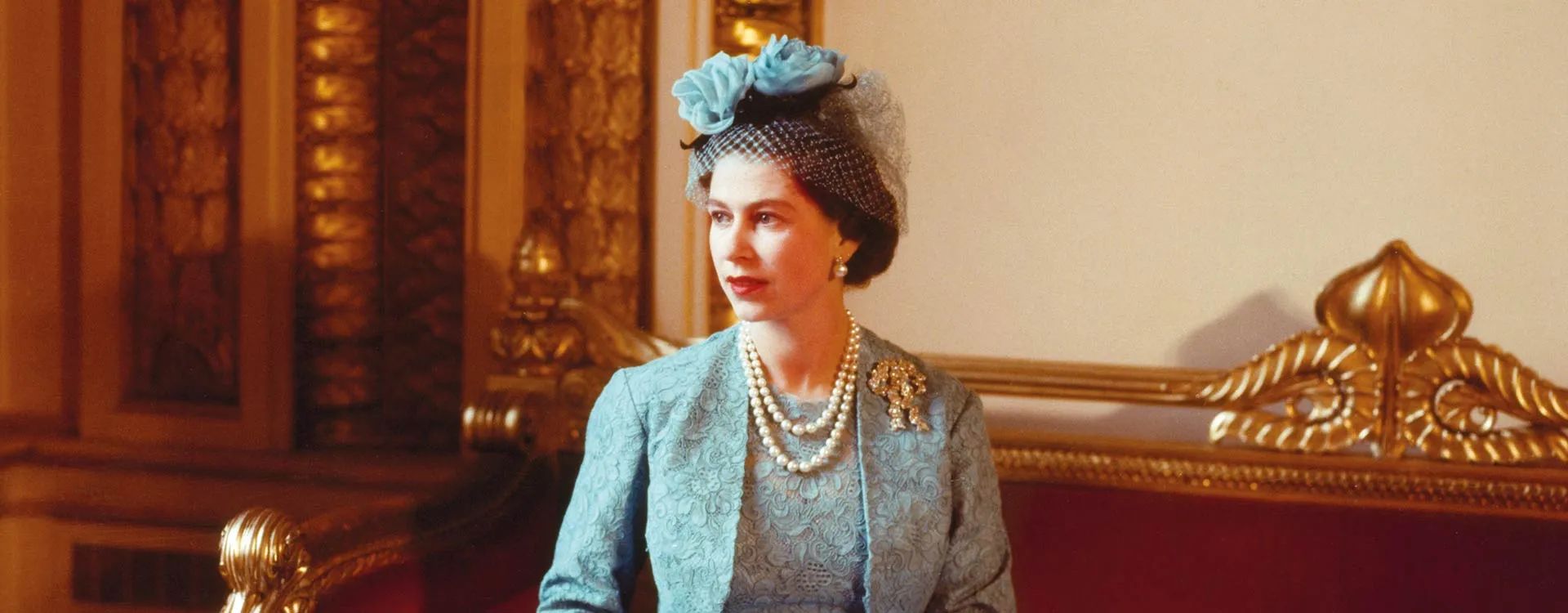 A woman in a blue lace dress and matching hat with a large flower sits on a red and gold ornate bench, wearing a pearl necklace and brooch—evoking the timeless elegance featured in Fashion Exhibitions, surrounded by opulent gold décor.