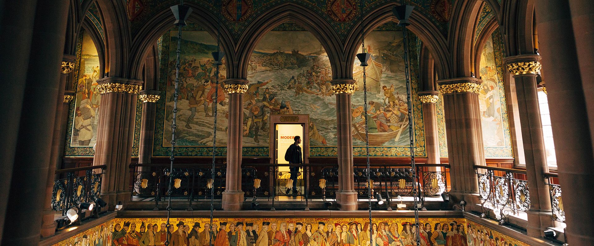 A person walks along a balcony inside a grand hall with ornate arches, elaborate wall murals depicting historical scenes, and intricate gold accents—an experience straight out of an Edinburgh travel guide packed with insider tips.