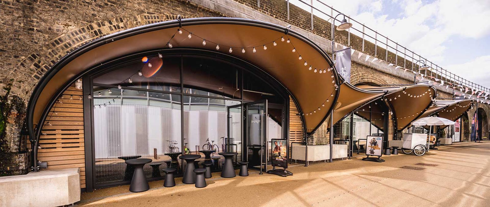 Curved brown awnings with fairy lights hang over a modern café exterior set against an arched brick railway viaduct, evoking the urban charm of London independent cinemas. Black tables and chairs are arranged outside along a wide paved walkway.