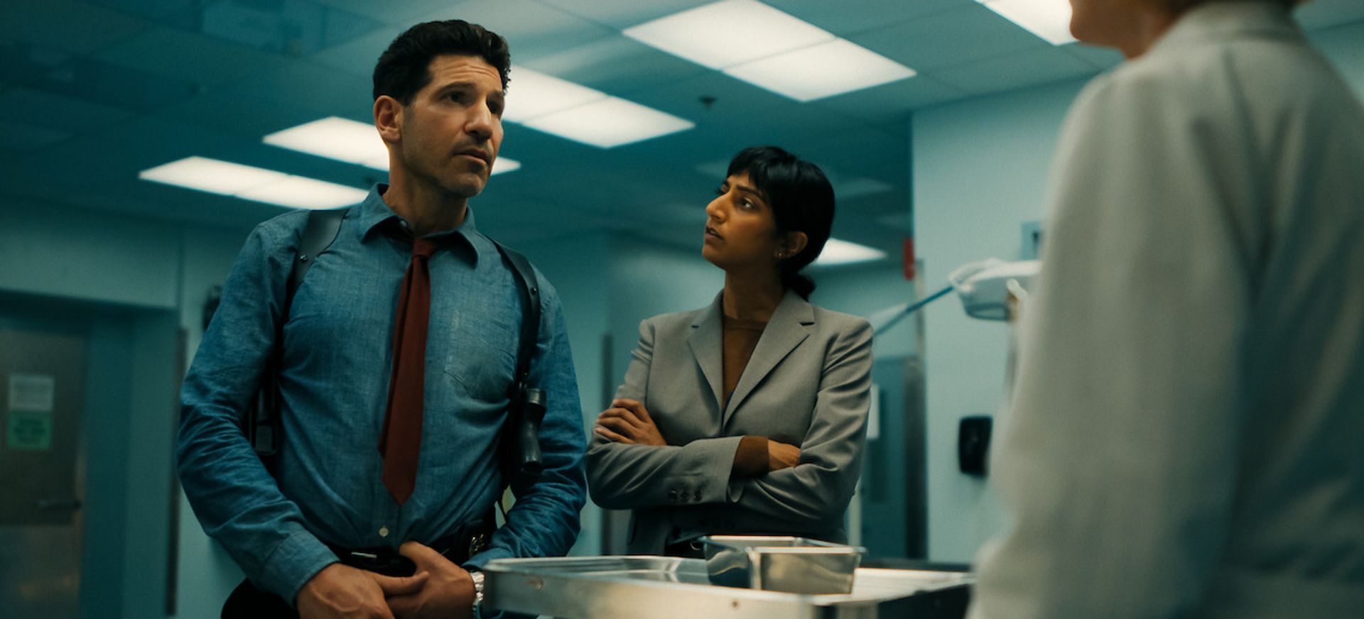 Two people in business attire stand in a brightly lit, clinical corridor, speaking with a third person whose back is to the camera. The man looks serious, while the woman stands with arms folded, channelling the intensity of a Netflix thriller.