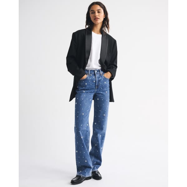 A woman stands against a plain background wearing a black blazer over a white shirt, blue high-waisted embellished jeans with small white embroidered designs, and black shoes, with her hands in her pockets.