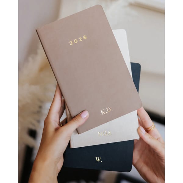 A person holds three 2026 diaries; the top is tan with 2026 and K.D. in gold, the middle is cream with NOA, and the bottom is black with W. Each diary has gold initials or letters on its cover.