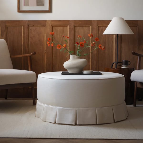 A round, cream-coloured ottoman with a pleated skirt stands in a living room, topped with a modern vase of orange flowers. Two beige armchairs and a cosy granny sofa flank it, with a lamp and wooden panelling in the background.