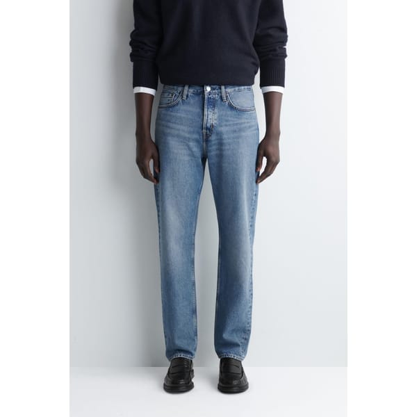 A person wearing a dark jumper, light blue straight-leg jeans, and black shoes stands facing forwards against a plain light background—showcasing effortless style tips for men. The photo is cropped at the shoulders.