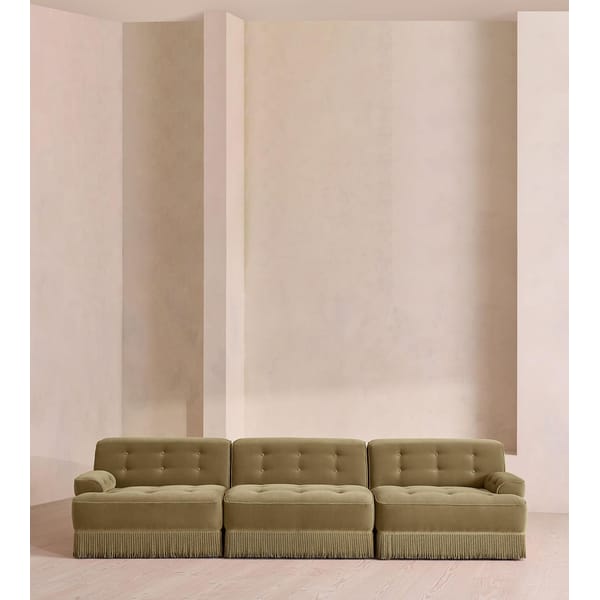 A beige, three-seater granny sofa with button tufting and fringed trim sits against a minimalist, light-coloured wall with geometric detailing—a modern take on vintage furniture that elevates home décor with its soft, neutral palette.