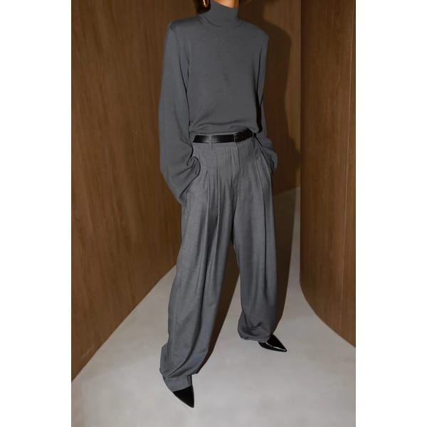 A person stands indoors wearing a loose-fitting dark grey polo neck, pleated trousers with wide legs, a black belt, and pointed black shoes. The setting features curved wooden walls and a light floor. The face is not visible.