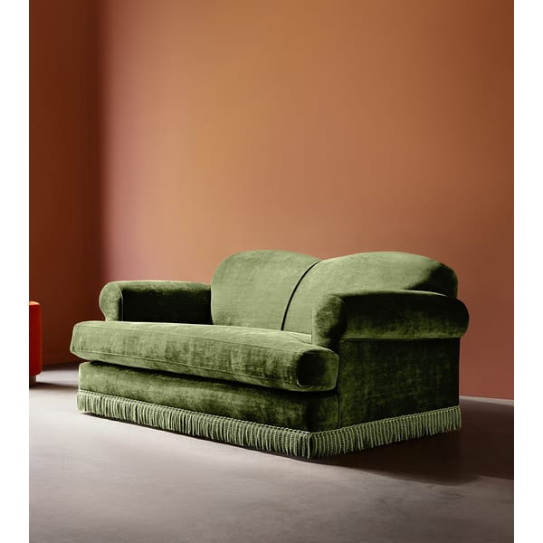 A plush green velvet granny sofa with rounded arms and decorative fringe stands against a smooth, warm terracotta-coloured wall on a grey floor, capturing the essence of vintage furniture style.