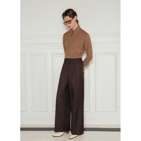 A person wearing a brown long-sleeved collared shirt, high-waisted wide-leg dark trousers, white shoes, and yellow-tinted sunglasses stands against a white panelled wall—offering style tips for men with this effortlessly cool look.
