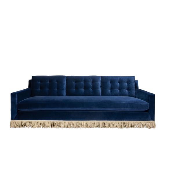 A deep blue velvet granny sofa with tufted back cushions and gold fringe trim along the bottom, set against a plain white background—perfect for elevating your home décor with timeless style.