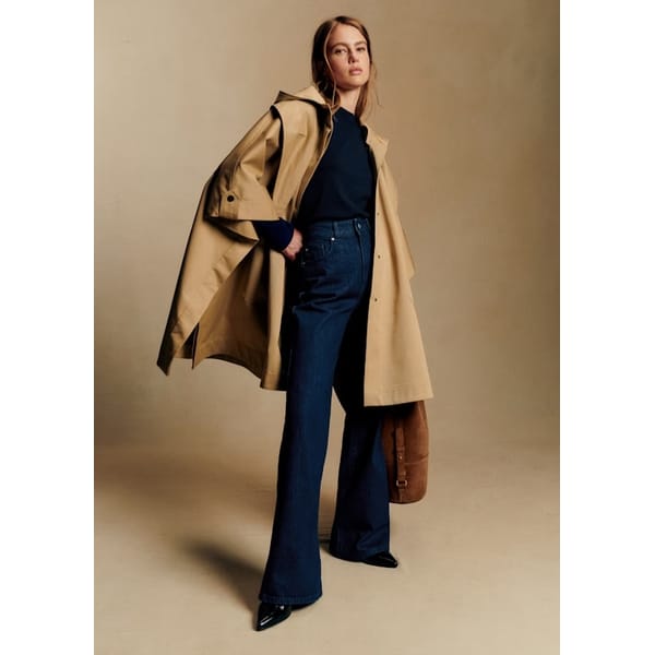A woman stands confidently in a tan trench coat, styled like one of the most stylish waterproof jackets, draped over her shoulders. She pairs it with a navy top, high-waisted flared jeans, black shoes, and a brown bag against a plain beige background.