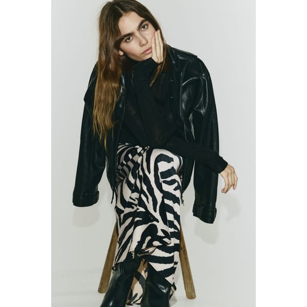 A person with long brown hair sits on a wooden stool, resting their head on their hand. Dressed in a black leather jacket and top, they wear striking zebra print trousers and black boots, set against a plain white background.