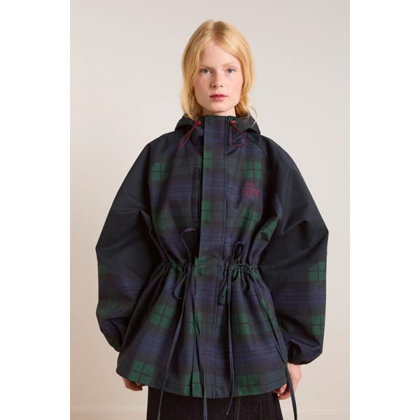 A young woman with blonde hair wears a stylish waterproof jacket—a dark checked hooded design with drawstrings at the waist and red embroidered detail on the chest—standing against a plain light background.