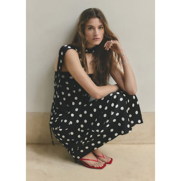 A woman with long brown hair squats against a cream wall, wearing a black polka dot dress perfect for wedding guest dresses, a matching scarf, silver bracelets, and red sandals. She looks slightly to the side with a neutral expression.