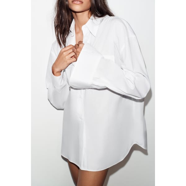 Channelling cool fashion inspired by Carolyn Bessette Kennedy, a person in an oversized white button-up shirt stands against a plain white background, hands near the collar and sleeves covering part of their hands, face partially out of frame.