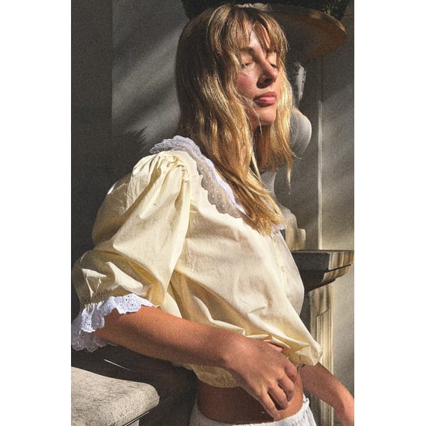 A young woman with long blonde hair and fringe, wearing a light yellow summer top with lace trim, stands in sunlight with her eyes closed, appearing relaxed and serene. Shadows and warm light highlight her face and outfit.