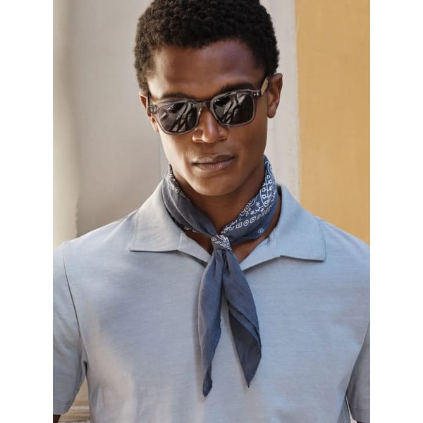 A man wearing dark sunglasses, a light grey polo shirt, and a blue patterned scarf tied around his neck showcases men's holiday fashion in front of a neutral background with a yellow accent wall.