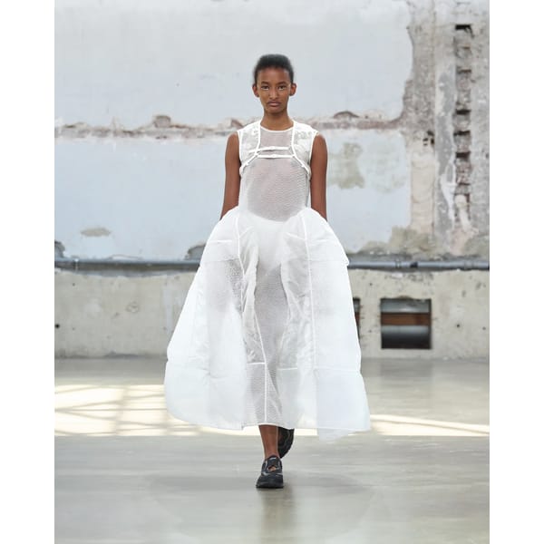 A model walks a catwalk wearing a sheer, textured white dress with voluminous sides and black shoes, set against an industrial concrete wall—offering edgy inspiration for unique Fathers Day gifts 2026.