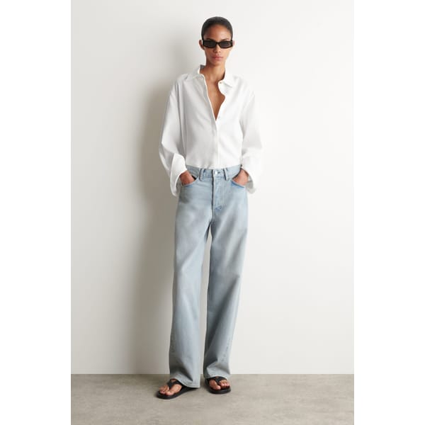 A person wearing a loose high street linen white button-up shirt, light blue baggy jeans, black sandals, and black sunglasses stands against a plain white wall with hands in their pockets.