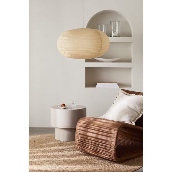 A cosy modern living space with a wavy wooden chair, a beige cushion, a round side table holding a glass of water and a snack, a woven rug, minimalist built-in shelves with decor, and a large pale paper ceiling light.