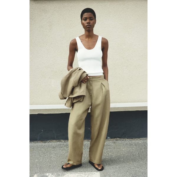 A person stands outdoors against a beige wall, wearing a white sleeveless top, high street linen wide-legged tan trousers, black sandals, and holding a tan jacket in one hand.