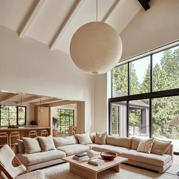 Bright, modern living room with large windows, neutral-toned corner sofa, wooden coffee table, textured rug, and a round paper ceiling light pendant. Forest scenery is visible through the windows.